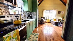 A kitchen with a stove, sink, and yellow kettle. There is a dining area with a table and chairs. The room has wooden floors and a high ceiling with exposed beams.