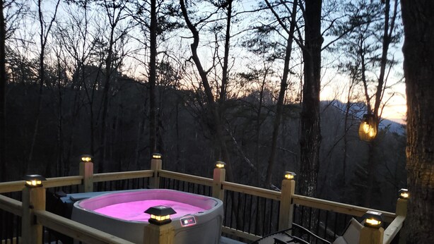 A hot tub on a wooden deck with railings and chairs, surrounded by trees.