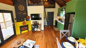 A spacious living room with a fireplace, wooden beams, a TV, and a kitchen with green cabinets and a stainless steel refrigerator.