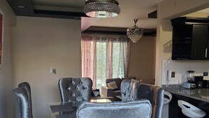 A modern dining area with a glass table and grey upholstered chairs. There is a chandelier above the table and a kitchen area with black cabinets and a countertop.