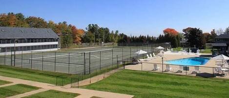 Cancha de tenis junto a una piscina con sillas de descanso y sombrillas.