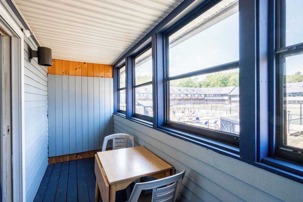 A small, enclosed balcony with a wooden table and two chairs, large windows offering a view of a parking area and buildings.