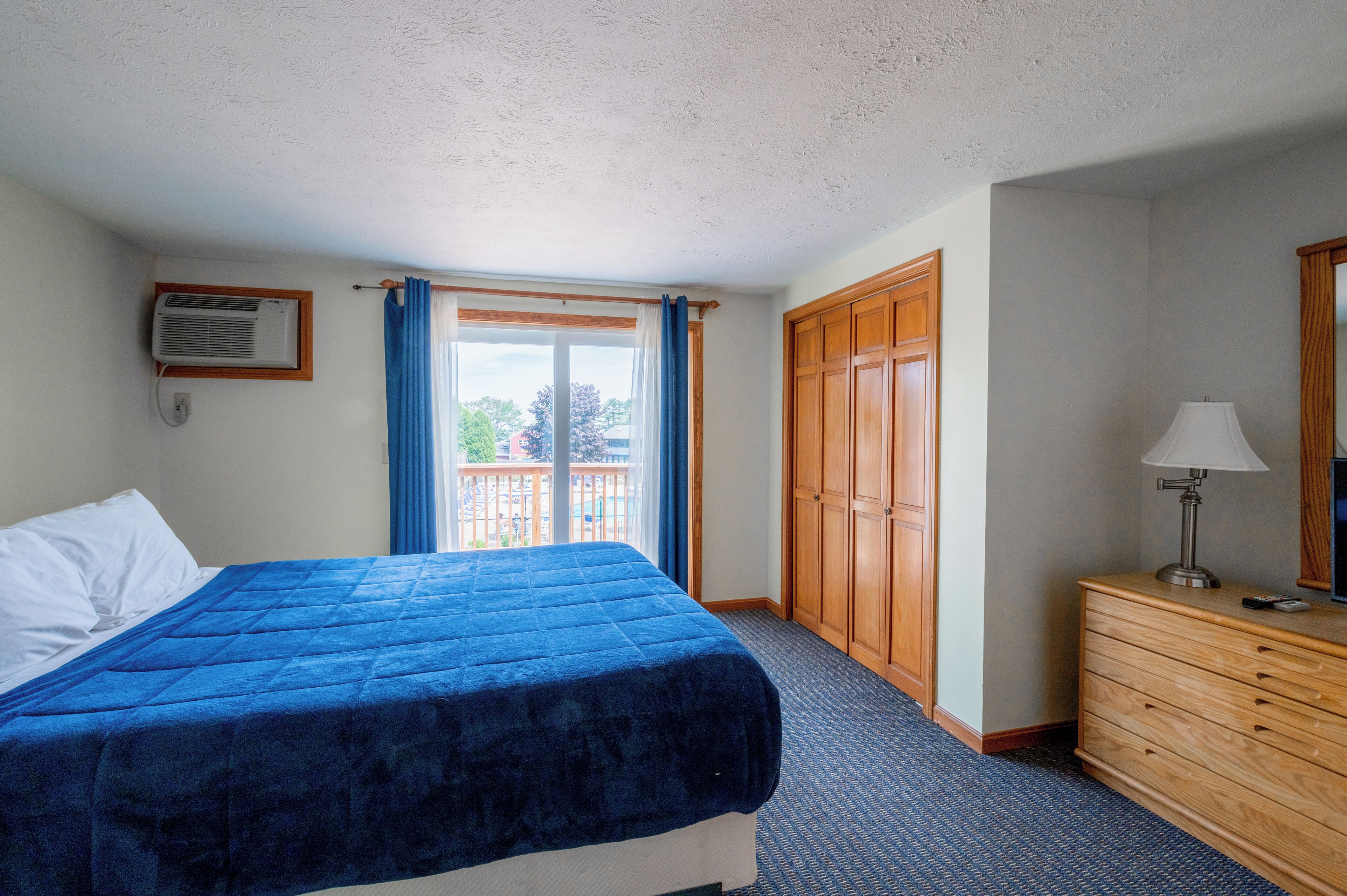 A hotel room with a bed, a dresser, a lamp, a window with curtains, and an air conditioning unit.