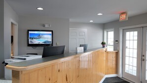 A reception desk with a flat-screen TV displaying a nautical theme, a wooden counter, and a reception area with a door and windows. - Nautical Mile (Wells)