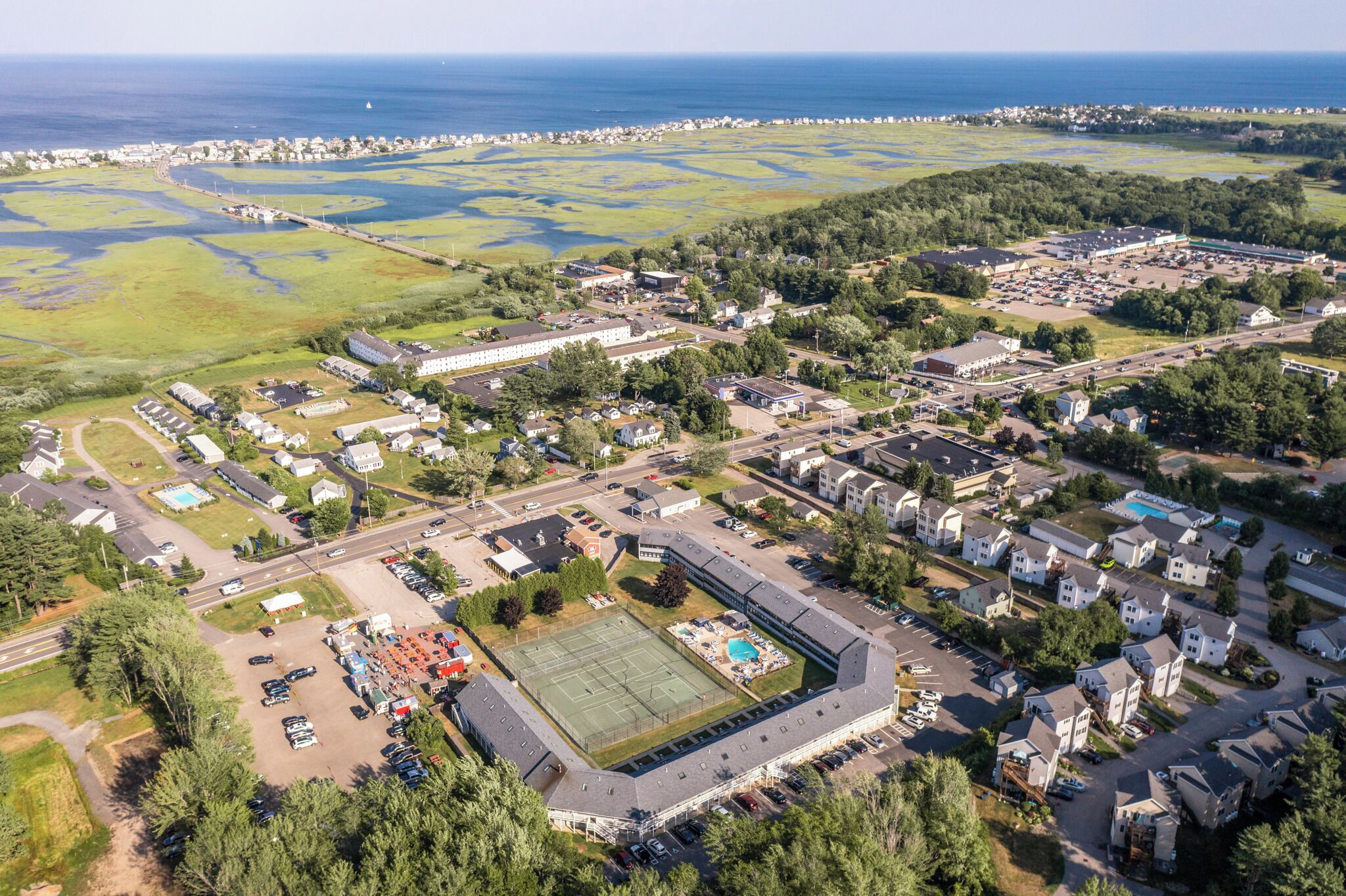 Eine Vogelperspektive eines Wohngebiets mit einem großen Sportplatz, mehreren Gebäuden und einem Gewässer im Hintergrund.