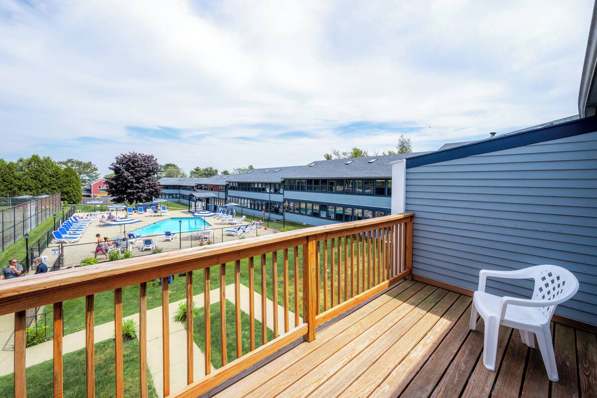 A balcony with a wooden railing overlooking a swimming pool area with lounge chairs and a large tree.