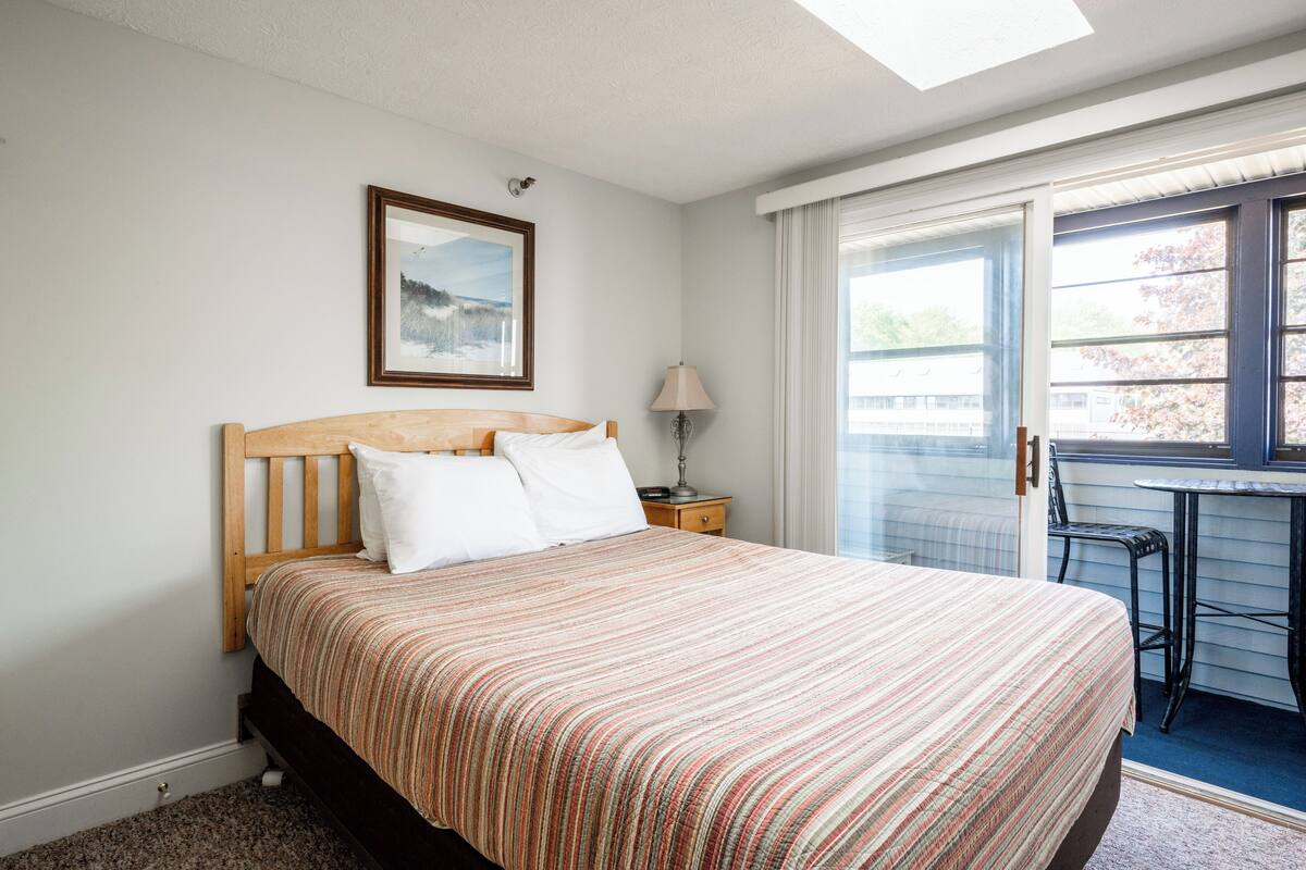 A bedroom with a bed, a nightstand, a lamp, a window, and a door leading to an outdoor area.