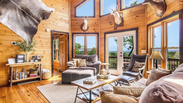 A cozy cabin interior with wooden walls, a fireplace, a sofa, a coffee table, and a large window overlooking the outdoors. - Family-Friendly Blue Ridge Log Cabin w Stunning Views-Free Flexible Check In-Out (Blue Ridge)