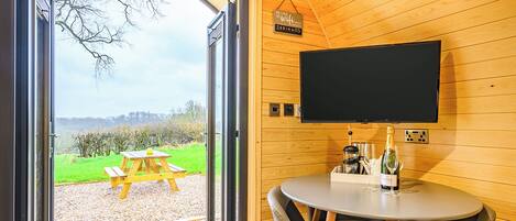 A modern room with a wooden interior, a large window, a flat-screen TV, a round table with chairs, and a picnic table outside.