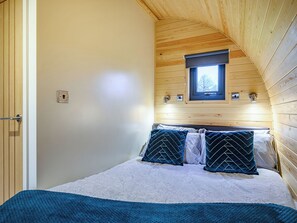 Une chambre avec un plafond en pente en bois, une petite fenêtre et un lit agrémenté d’oreillers à motifs.