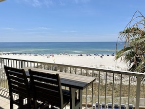 A beach view from a balcony with a table and chairs, overlooking a sandy beach with umbrellas and people.