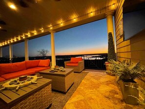 Área externa com mesas e cadeiras, vista para o pôr do sol, incluindo um sofá vermelho, duas poltronas, uma mesa de centro e plantas em vasos.