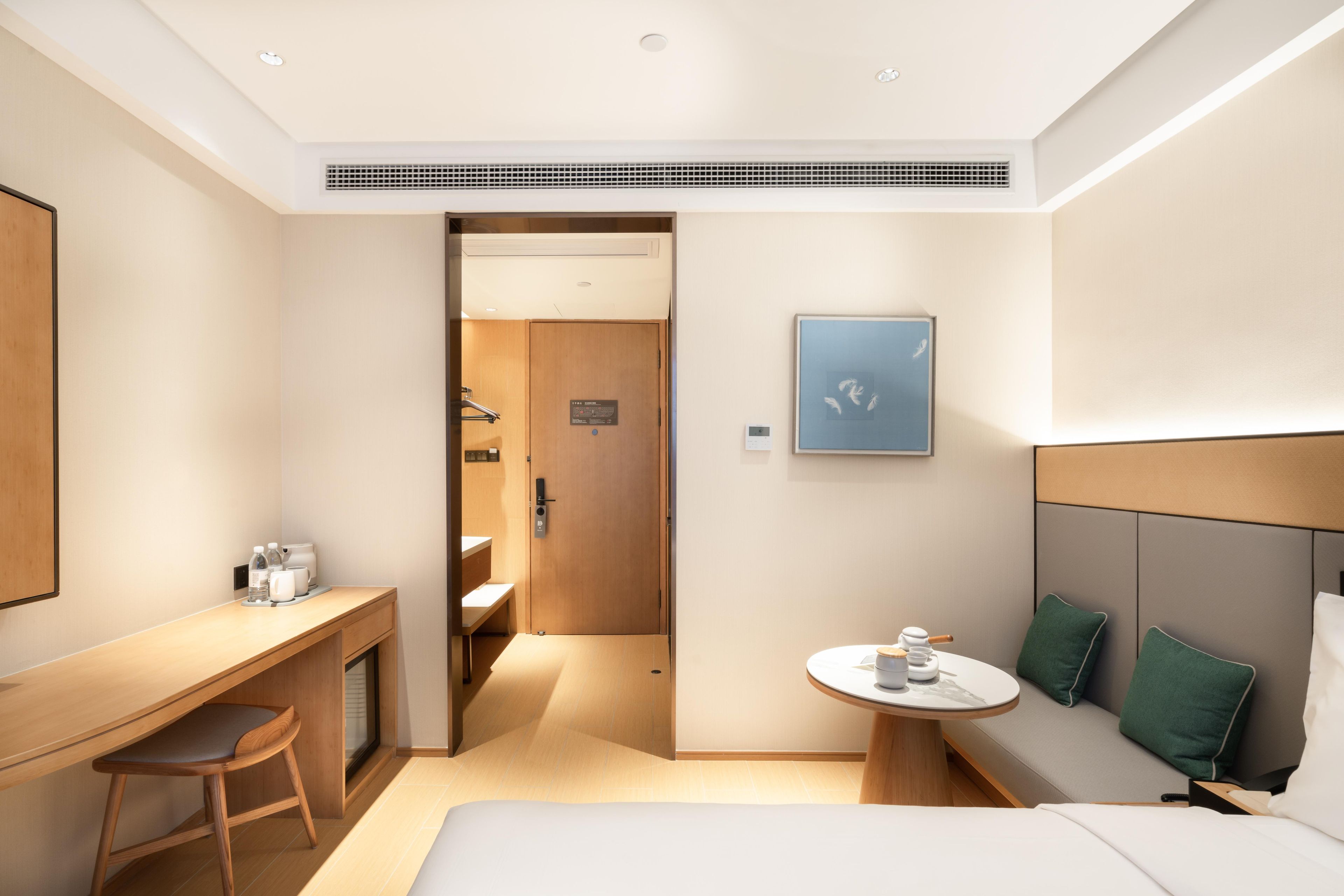 A modern hotel room with a bed, a desk, a small round table, and a bathroom visible through an open door.
