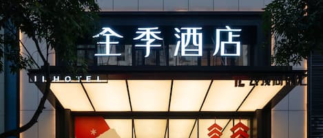 A hotel entrance with a sign in Chinese characters, a large illuminated window with festive decorations, and a partial view of the building's exterior.