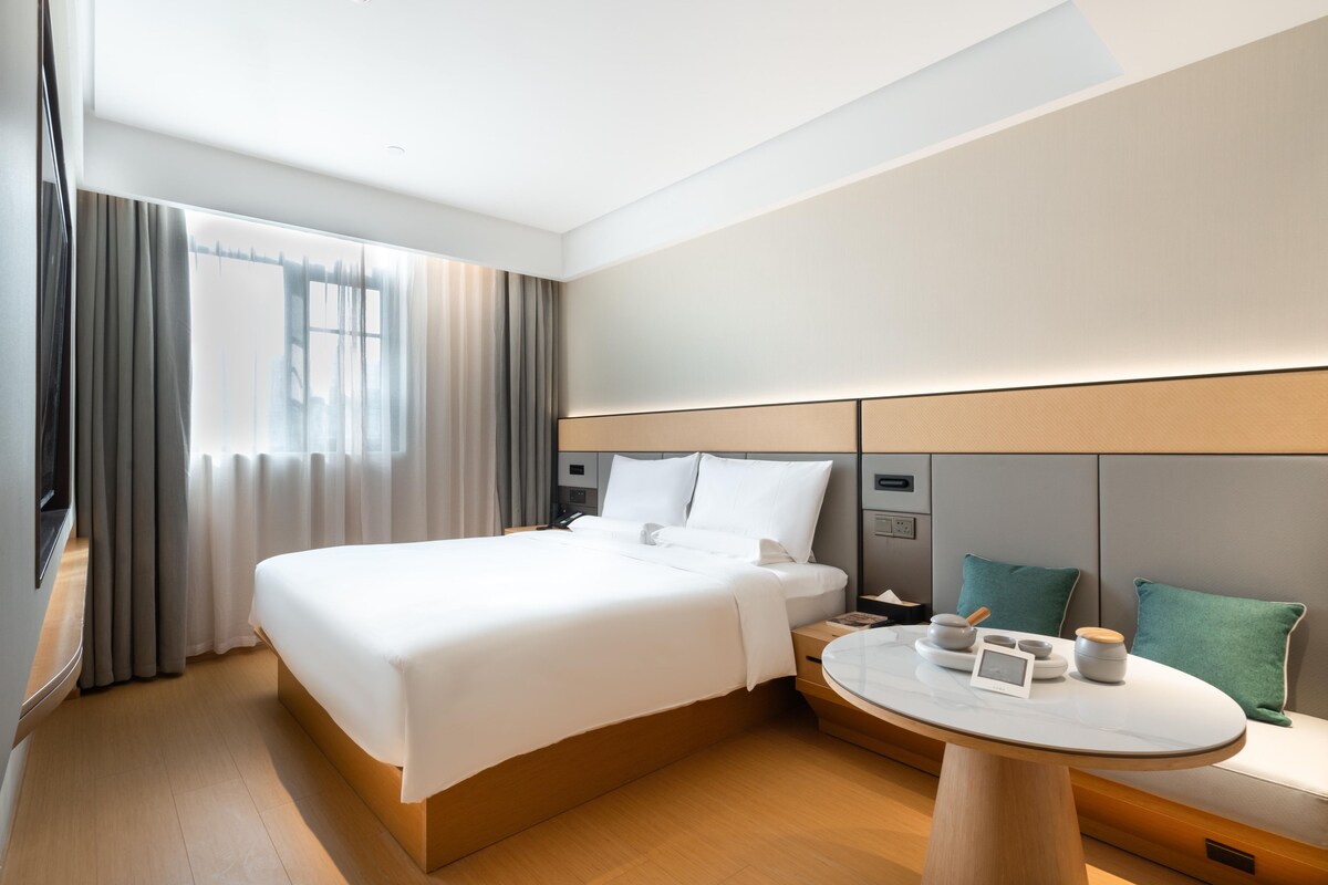 Une chambre d’hôtel avec un grand lit, des tables de chevet, une table ronde avec une lampe et une vue sur l’extérieur.