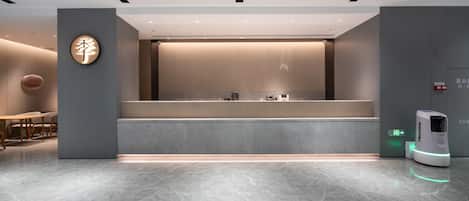 Een moderne hotelontvangsthal met een marmeren vloer, een receptiebalie, een digitale informatiezuil en een zithoek met een ronde tafel en stoelen.