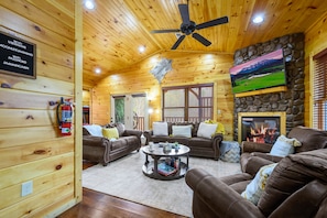 A cozy cabin interior with wooden walls, a ceiling fan, a stone fireplace, and a television mounted on the wall.