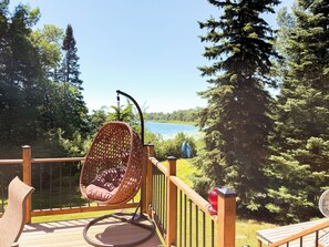 Eine Holzterrasse mit einem Korbstuhl, der auf einen See blickt, umgeben von Bäumen.