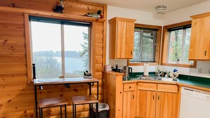 Une cuisine rustique aménagée dans une cabane en bois, avec une grande fenêtre, des placards en bois, un évier et une cuisinière.