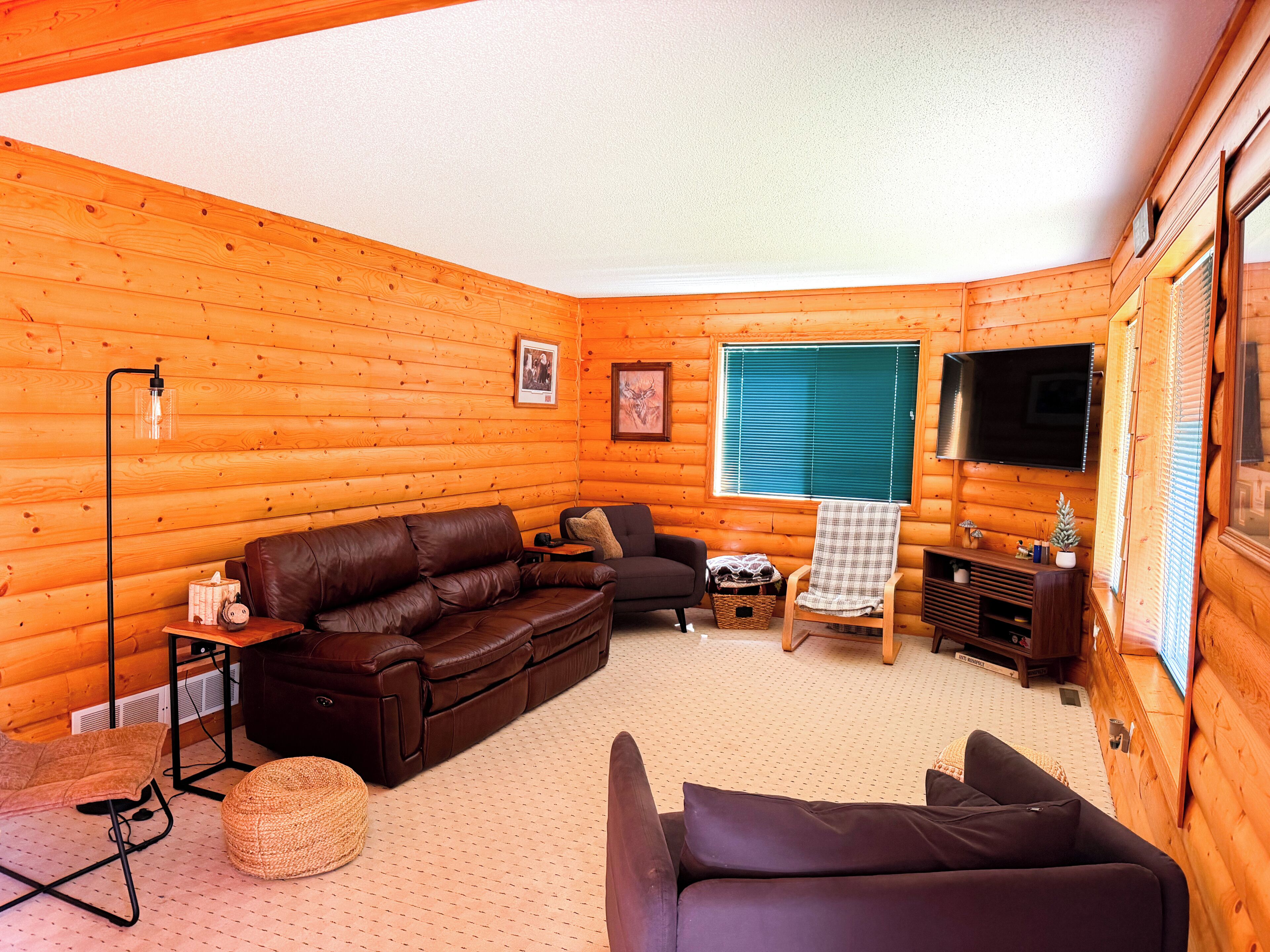 Interior de una cabaña de troncos con un sofá de cuero marrón, un sillón más pequeño, un televisor y una ventana con persianas.