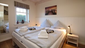 A bedroom with two beds, a nightstand, a lamp, a window with curtains, a painting on the wall, and a mirror in the adjacent room. - Comfortable 5-star vacation home with approx. 55 sqm living space on the first floor (Wyk auf Foehr)