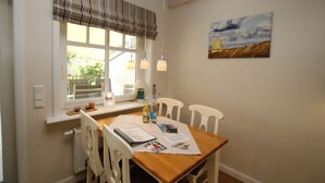 A dining area with a wooden table, white chairs, a window with a striped valance, and a painting on the wall. - Comfortable 5-star vacation home with approx. 55 sqm living space on the first floor (Wyk auf Foehr)
