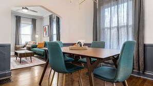 A modern dining area with a wooden table and teal chairs, a sofa with orange cushions, and a large window with curtains.