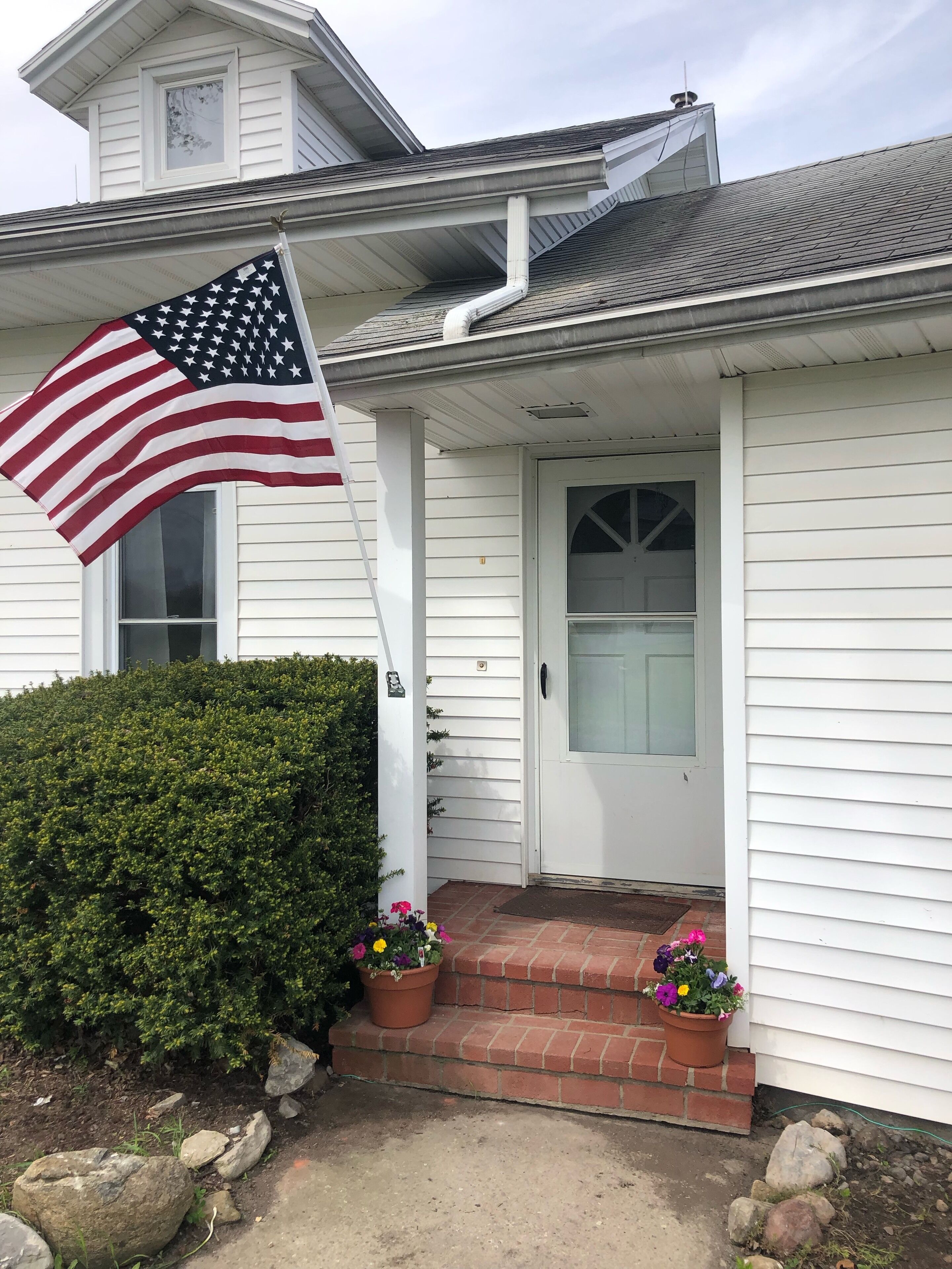 Une maison blanche avec un porche à l’avant, un drapeau et des plantes en pot.