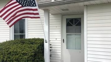 Une maison blanche avec un porche à l’avant, un drapeau et des plantes en pot.