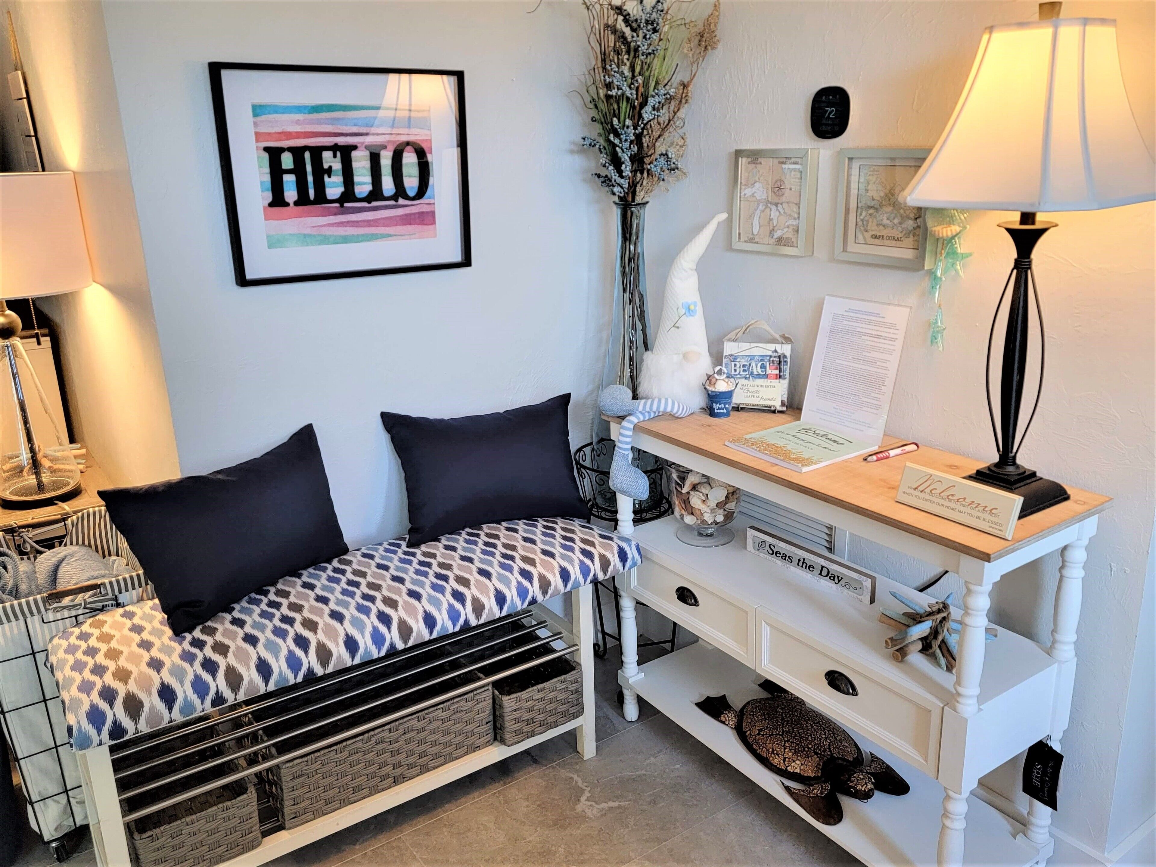 A cozy room with a patterned bench, a desk with a lamp, a framed 'HELLO' sign, and a vase with dried flowers.