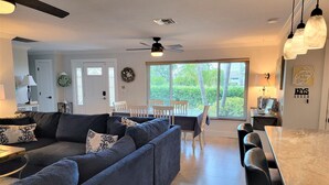 A modern living room with a sectional sofa, dining table, and chairs. There is a ceiling fan, large windows with blinds, and a view of greenery outside.