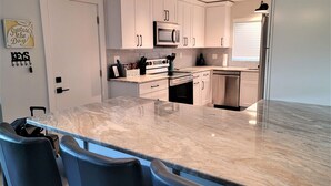 A modern kitchen with white cabinets, a large marble countertop, and built-in appliances. There are three black bar stools in front of the counter.