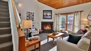 A cozy living room with a fireplace, wooden ceiling, and a staircase.