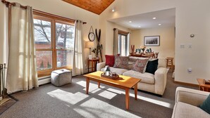 A cozy living room with a wooden ceiling, large windows with curtains, a sofa, a coffee table, and a fireplace.