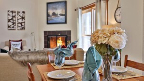 A dining area with a table set for two, a vase with flowers, and a fireplace in the background.