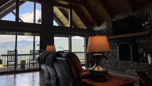 Ein geräumiges Zimmer mit hoher Decke, großen Fenstern mit Blick auf die Berge, einem Ledersofa, einem Tisch mit Lampe und einem Kamin aus Stein.