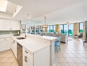 A modern kitchen with a central island, a dining area with chairs, a sofa, and a view of the ocean through large windows.