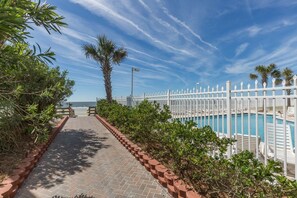 A pathway leading to a pool area with a white fence, palm trees, and a clear blue sky.