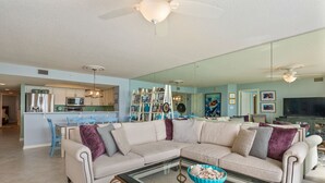 A living room with a large sectional sofa, a glass coffee table, and a kitchen area in the background.