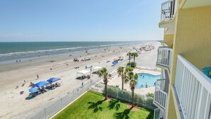 A beach view from a balcony overlooking a sandy beach with umbrellas, lounge chairs, and a pool.