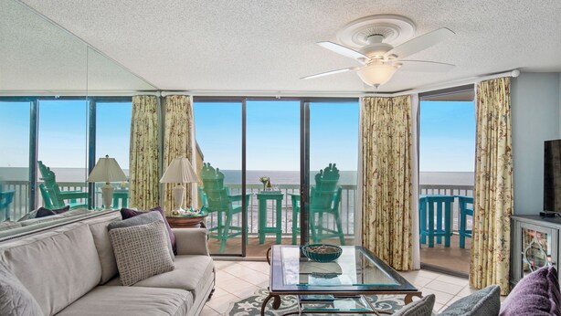 A living room with a sofa, coffee table, and chairs, a ceiling fan, and large windows with curtains.