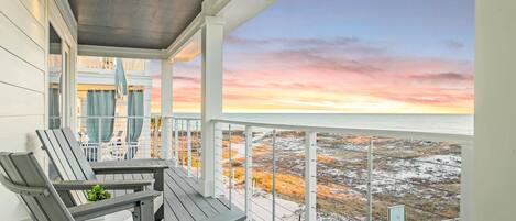 A wooden deck with two chairs and a small table, overlooking a beach with a sunset view.