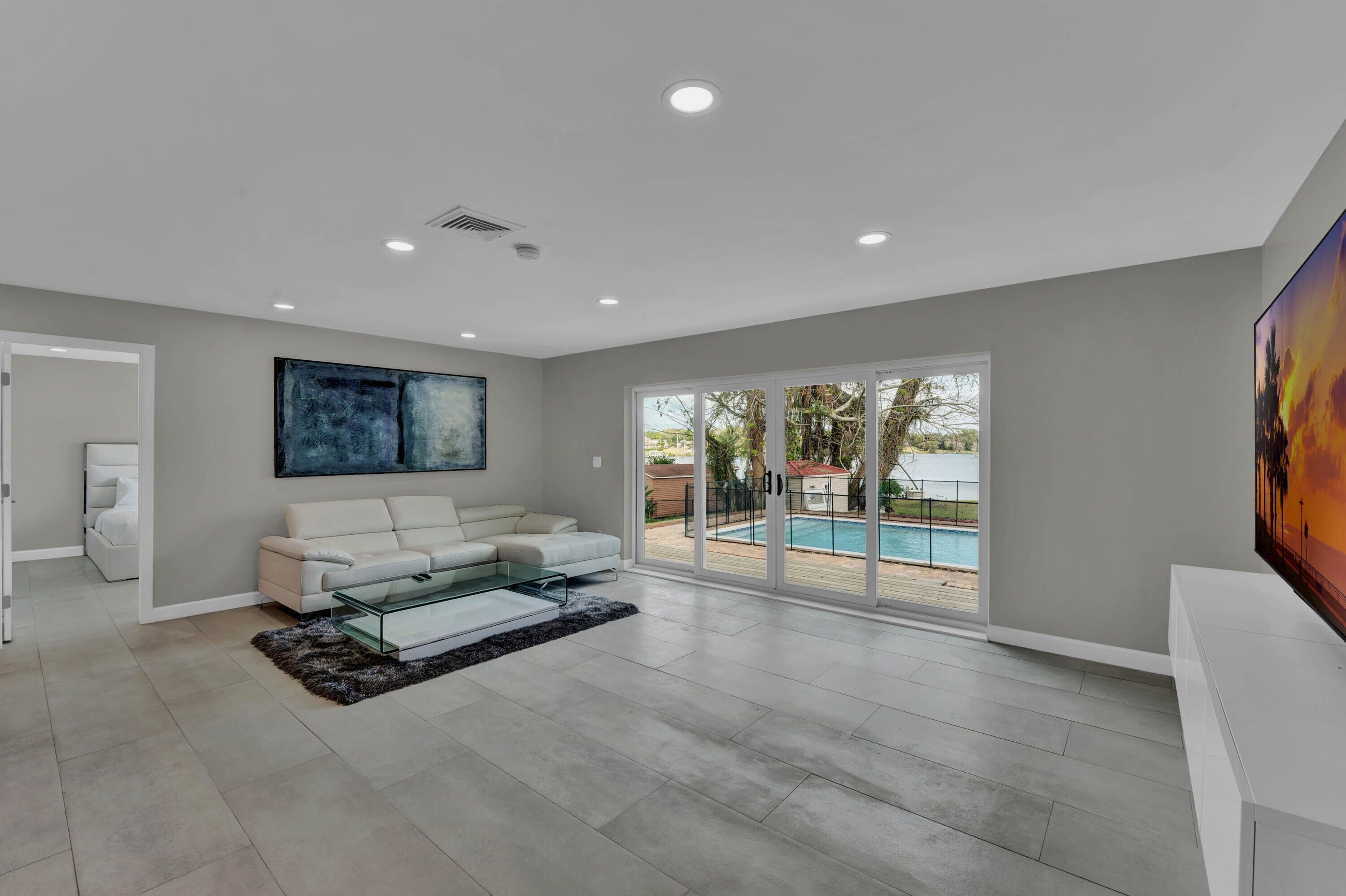 A spacious living room with a white sofa, a glass coffee table, and a large painting. There is a sliding glass door leading to a pool area.