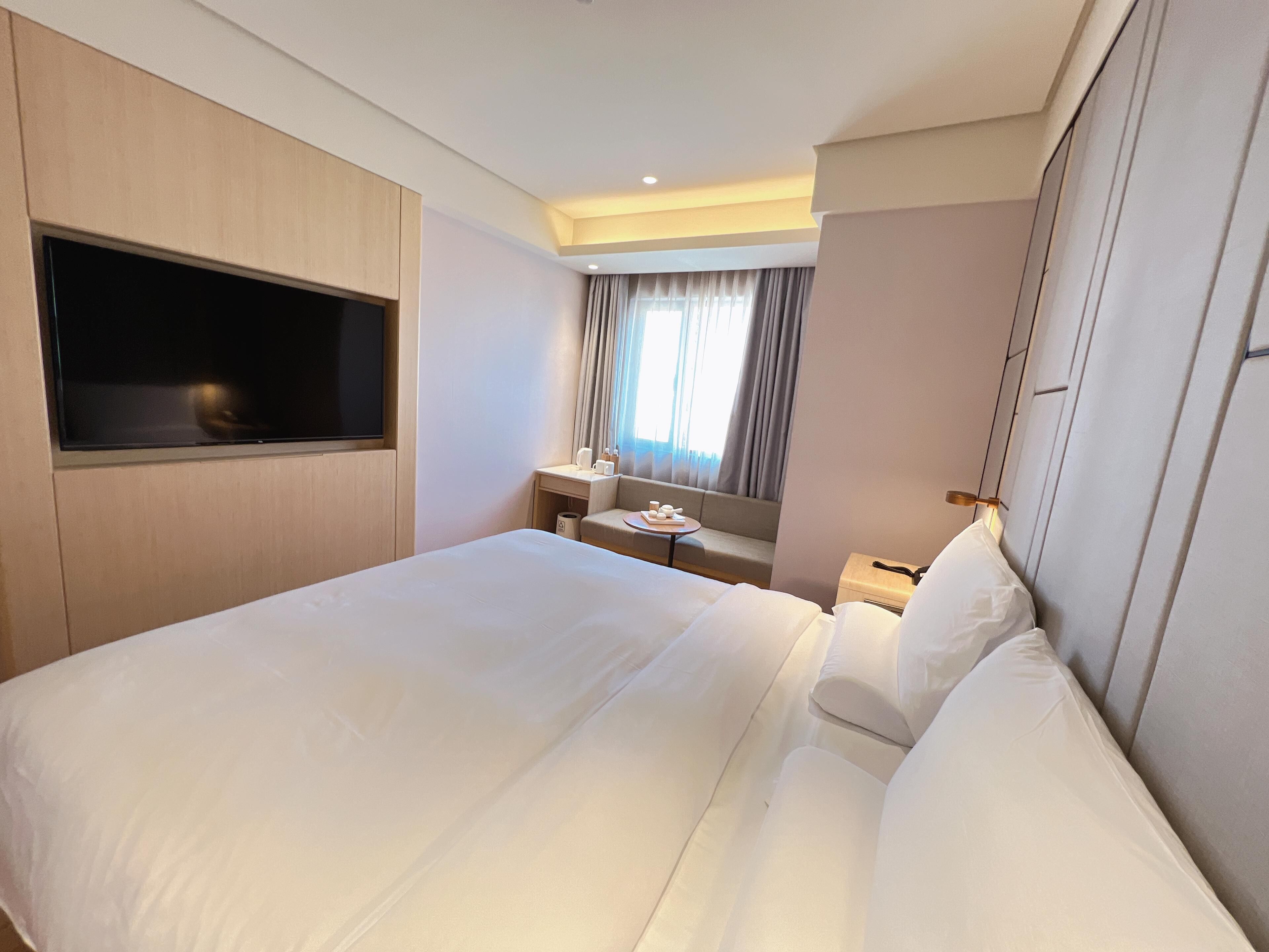 Una habitación de hotel con una cama grande, un televisor instalado en la pared, una mesita con lámpara y una ventana con cortinas.