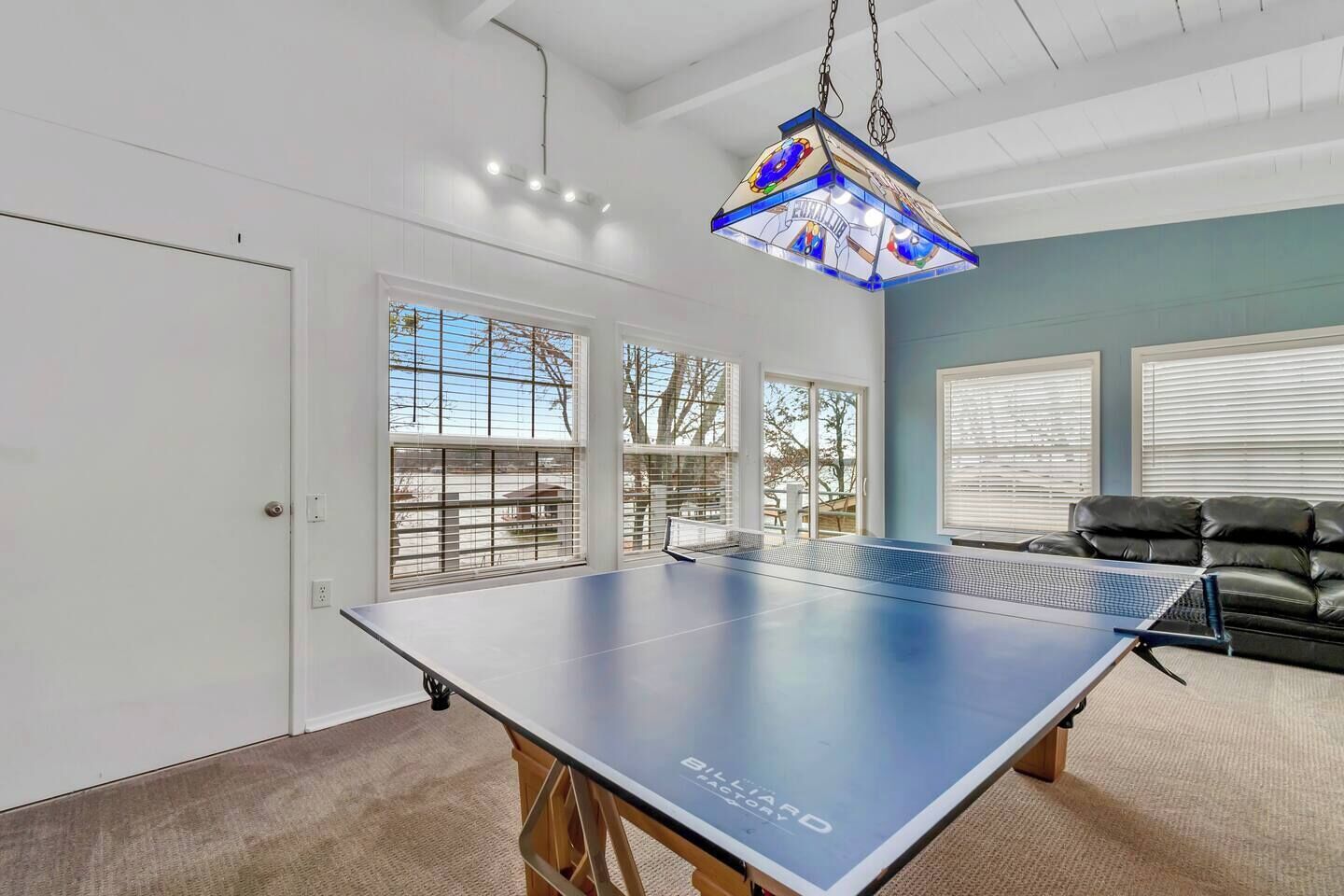 Une table de ping-pong dans une pièce dotée de grandes fenêtres, d’un luminaire au plafond et d’un canapé en cuir noir.