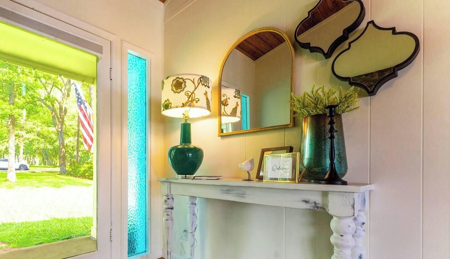 Quarto com uma mesa consola branca, um abajur verde, um espelho e um vaso com folhagem.
