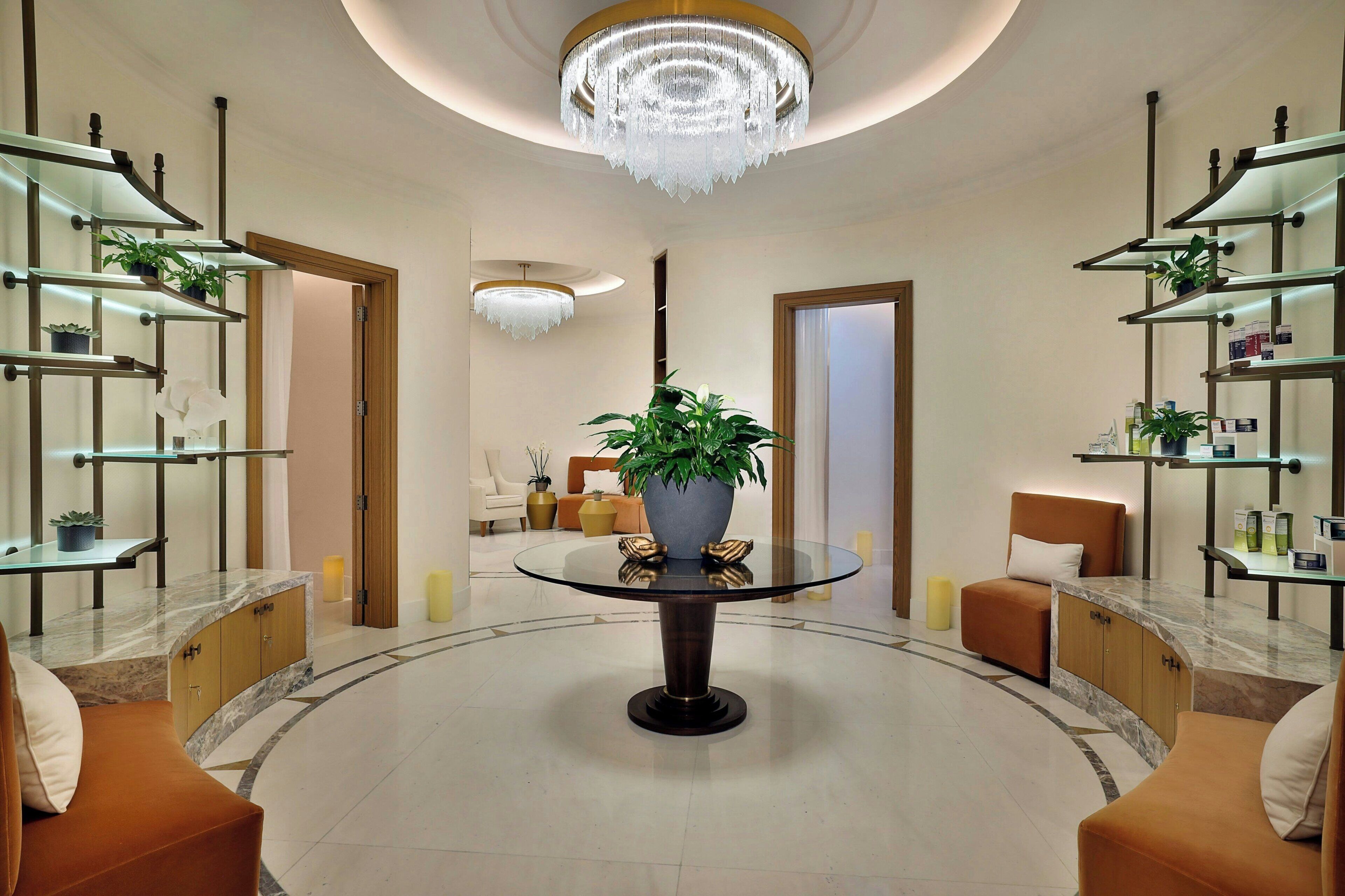 A lobby with a central glass table, surrounded by seating, and shelves with plants and items on either side.