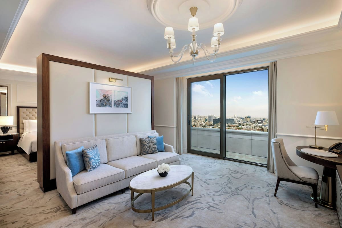 Une chambre d’hôtel moderne comprenant un canapé, une table basse, un fauteuil, un bureau avec une lampe, une grande fenêtre donnant sur la ville et un lustre.