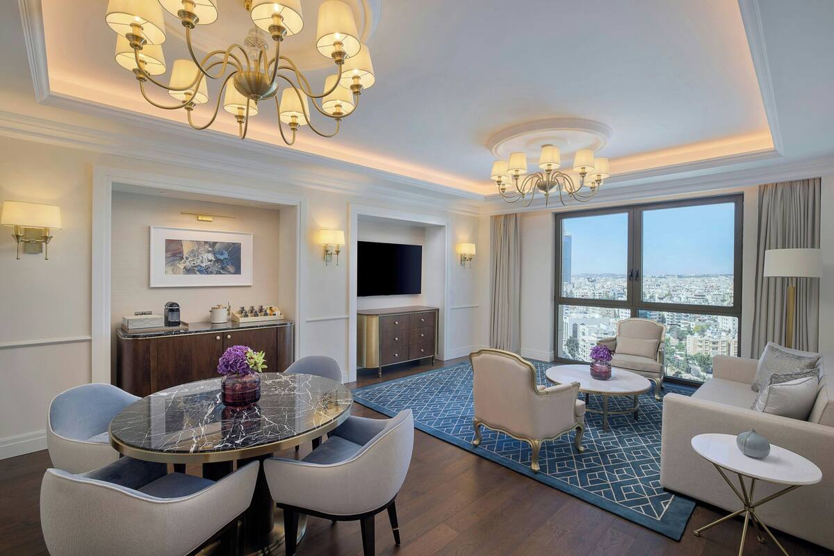 Une chambre d’hôtel comprenant un coin repas, un ensemble de canapés, une télévision et une grande fenêtre donnant sur la ville.
