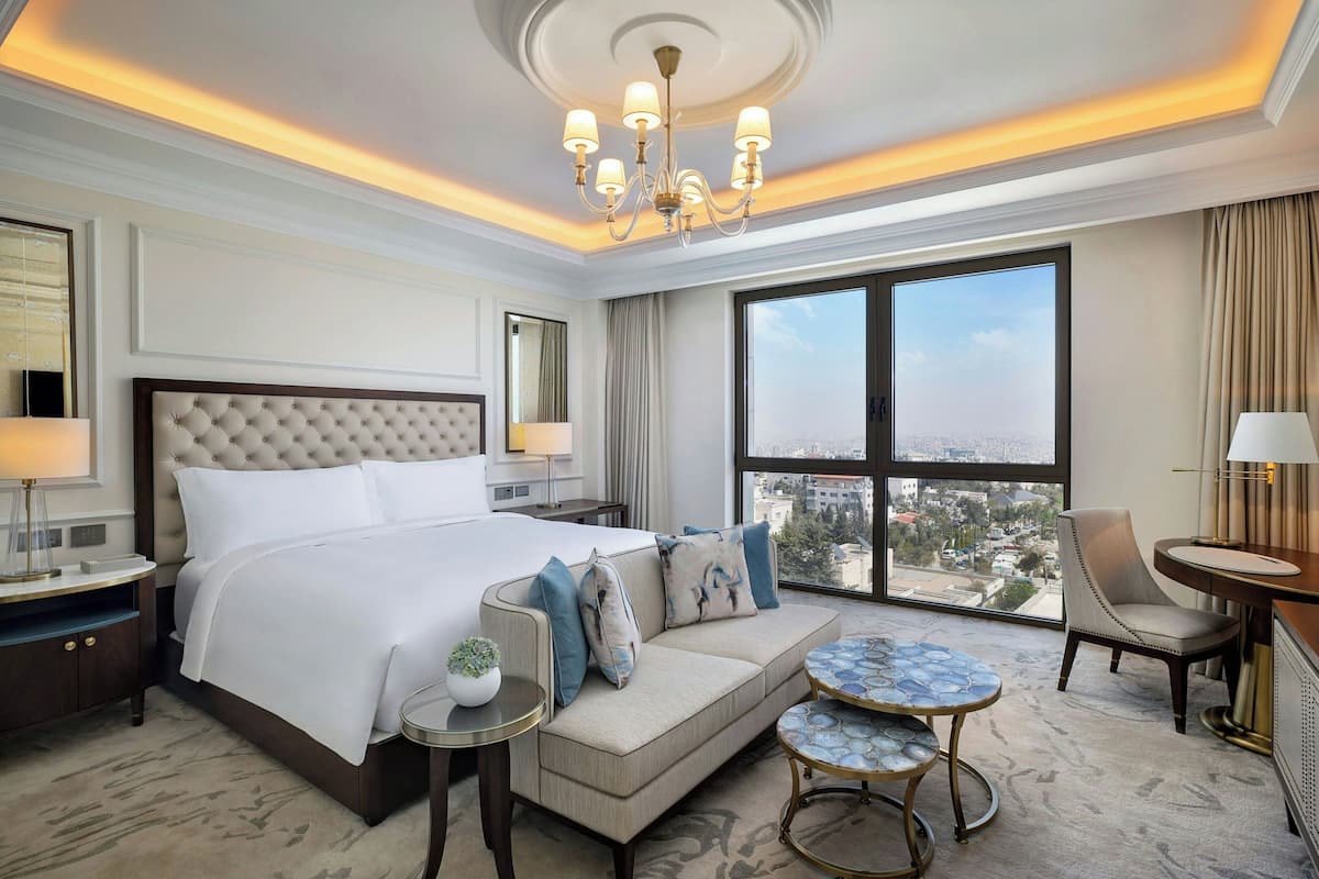 Une chambre d’hôtel avec un grand lit, un canapé, une petite table ronde, une chaise et une vue sur la ville grâce à de grandes fenêtres.